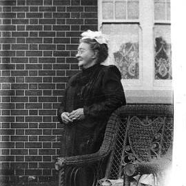 An unidentified elderly woman standing alongside a wicker chair.