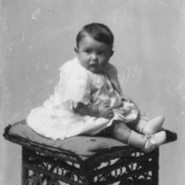 Portrait of an unidentified young child seated on a wicker stool.
