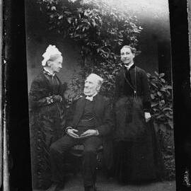 A seated elderly man flanked by two standing women.