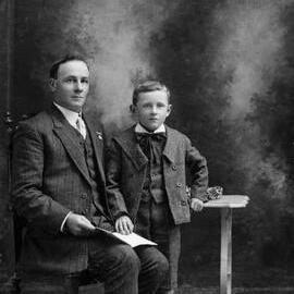 Studio portrait of a boy standing beside a seated man.  Family name Maher.