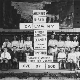 A group of children and young women on a stage with a large cross displaying a Christian message.