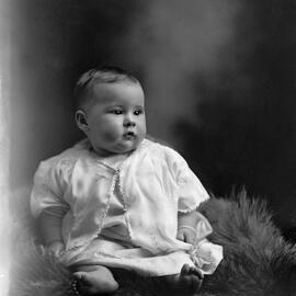 Studio portrait of a baby seated on a fur rug.  Family name Rowell or Howell.