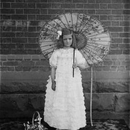A girl in a long dress standing outdoors on a rug in front of a wall.  Family name possibly Knee.