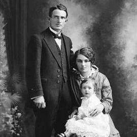 Studio portrait of woman holding a baby and seated alongside a man.  Family name Holmes.