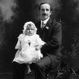 Studio portrait of a seated man holding a bare-footed baby.  Family name Walsh.