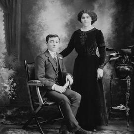 Studio portrait of a young woman standing alongside a seated young man.  Family name possibly Hartley.