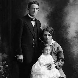 Studio portrait of woman holding a baby and seated alongside a man.  Family name Holmes.