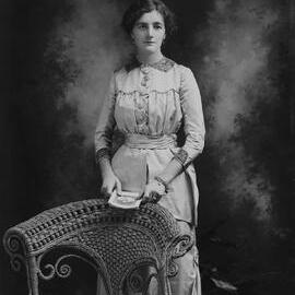 Studio portrait of a woman standing in front of a wicker chair.  Possible name Miss Stebbing.