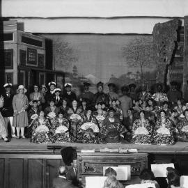 A large group of performers on a stage, some dressed as geishas.