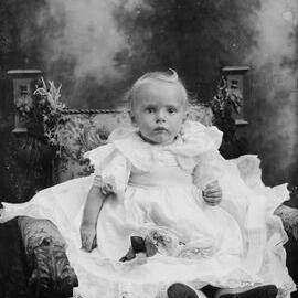 Studio portrait of a baby seated in an elaborate chair.