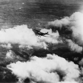 An aerial view of a single engine biplane flying above cloud and unidentified countryside.