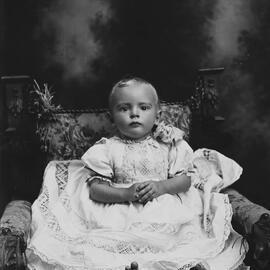 Studio portrait of a baby wearing an elaborate dress.  Family name possibly Lang.