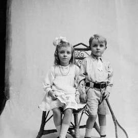 Studio portrait of a girl and a boy, seated together on wicker chair.  Family name Mc Lean.