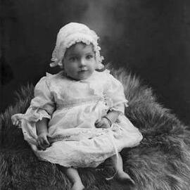 Studio portrait of a baby seated on a fur rug.   Family name Bell.