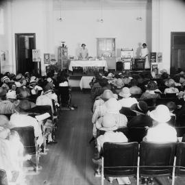 A gas appliance and cooking demonstration in a hall filled with women.