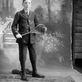 Studio portrait of a young suited man holding a stringed racquet.  Family name Corrigan.