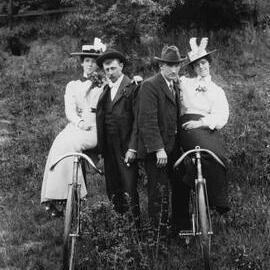 Two women seated on bicycles supported by two men.