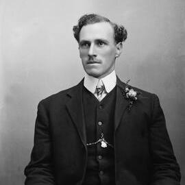 Studio portrait of a man wearing a button-hole spray of flowers.  Family name possibly Stewart.