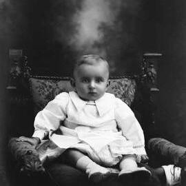 Studio portrait of a baby seated on a cushion.   Family name Drake.