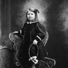 Studio portrait of a young girl standing on a wicker chair.  Family name Rawlings.