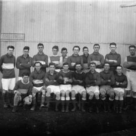 A group of men in football attire posed in front of a corrugated iron building.