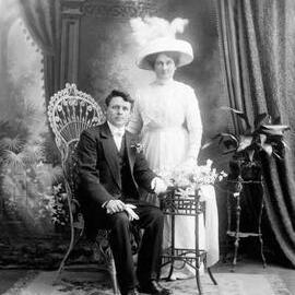 Studio portrait of a seated man and a standing woman holding flowers.  Family name Mc Coy.