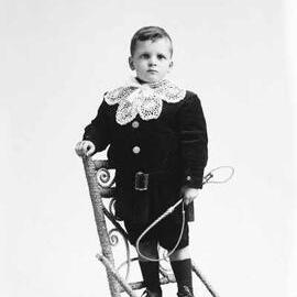 Studio portrait of a boy standing on a high-backed wicker chair.  Family name Randell.