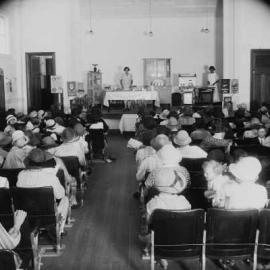 A gas appliance and cooking demonstration in a hall filled with women.