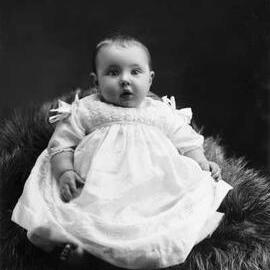Studio portrait of a baby seated on a fur rug.   Family name Daly.