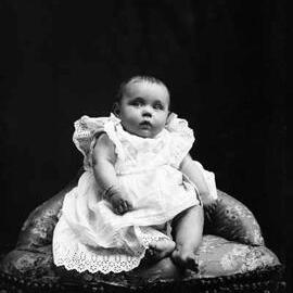 Studio portrait of a baby wearing a dress and seated on a corner chair.