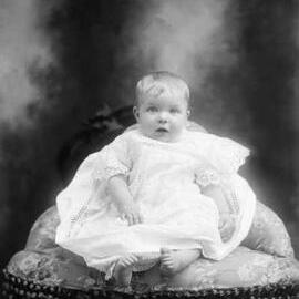 Studio portrait of a baby seated on an upholstered corner chair. Family name West.