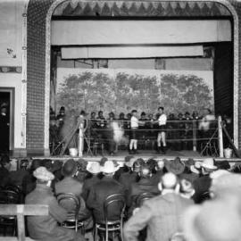 A boxing match on the stage of a hall with a male audience in attendance.