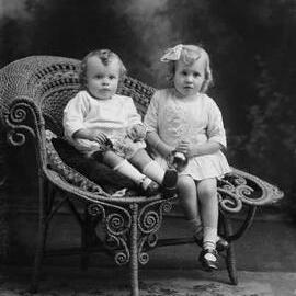 Studio portrait of a baby and a girl seated together on wicker chair.  Family name Carle.