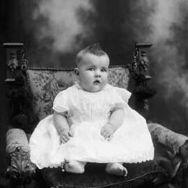 Studio portrait of a baby seated on a cushion and a carved chair.  Family name Carroll.