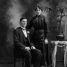 Studio portrait of a  woman wearing a black dress and a man wearing a suit and bow tie.  Family name Trethowan.