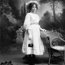 Studio portrait of a young woman standing at a wicker chair.  Family name possibly Leicester.