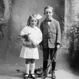 Studio portrait of a boy holding a stringed racquet and girl holding a large lily.   Family name Taylor.