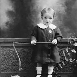 Studio portrait of an infant holding a riding crop.  Family name Petersen.