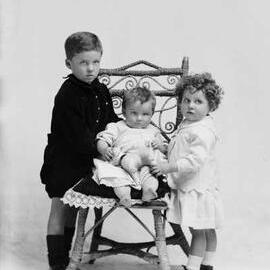 Studio portrait of a boy and girl standing alongside a seated baby.  Family name Lucas.