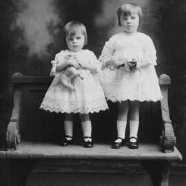 Studio portrait of two young girls standing on a garden seat.   Family name Groves.