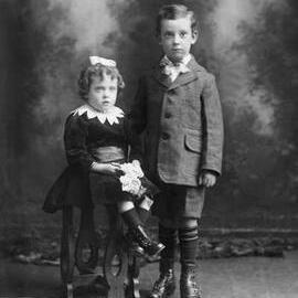 Studio portrait of a boy standing alongside a seated girl.   Family name Dowd.