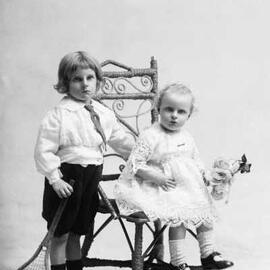 Studio portrait of  a young boy standing alongside a seated baby girl .   Family name Dynon.
