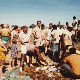 Malcolm Fraser in the Northern Territory standing with others gathered around food being cooked in an earth oven.