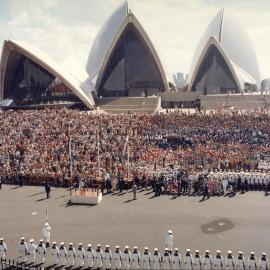 Queen Elizabeth II standing on a dais on the forecourt of the Sydney Opera House after her arrival in Sydney as part of her 1977 official Australian visit.