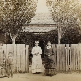Unidentified women and children in front of a house and garden at an unknown location.