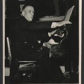 Francis Poulenc in a suit, sitting at a piano with his arms outstretched
