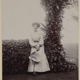 [Woman holding hat posing in front of hedge]