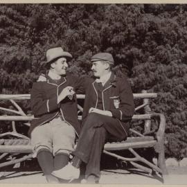 [Two men sitting on bench with log frame]