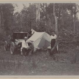 [Seated man and woman standing next to tent and motor car]