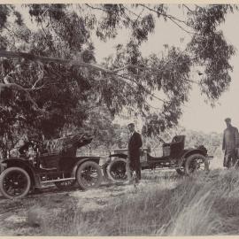 [Two men standing next to two motor vehicles]